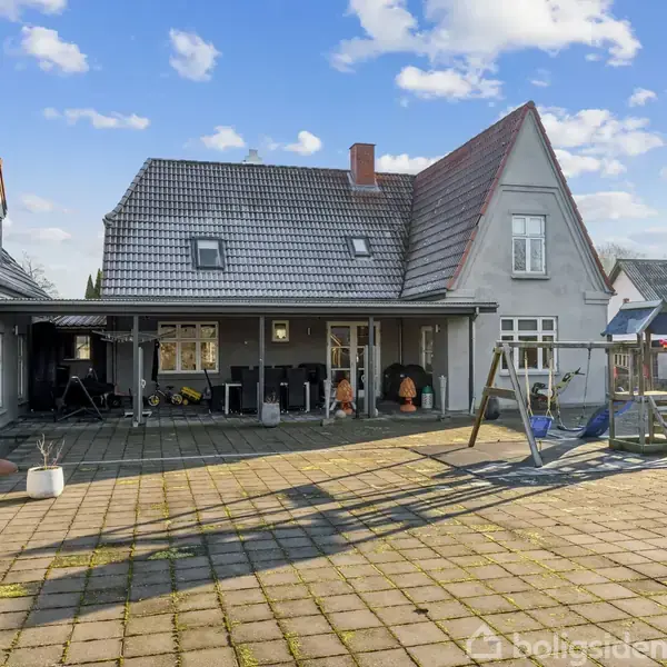Et gråmalet hus med saddeltag står i en gårdhave med fliser. Der er en overdækket terrasse og en gynge i forgrunden. Himlen er blå med få skyer.