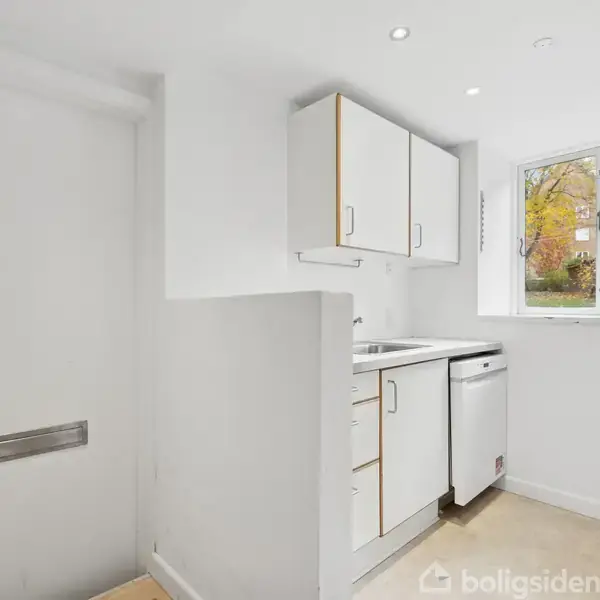 Køkken med hvide skabe og stålvaske ved vindue. Opvaskemaskine under bordpladen. Lyse vægge og gulv skaber et enkelt udseende. Dør til venstre, indendørs miljø.