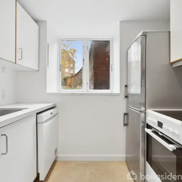 Køleskab står ved siden af en moderne ovn og skuffer i et kompakt, hvidt køkken. Et vindue i venstre side lader naturligt lys ind.