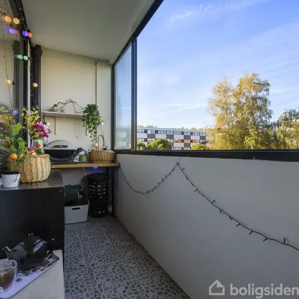 En altan dekoreret med planter og lyskæder ligger i en bymæssig boligblok. Terrassedøren står på klem, og udsigten viser træer og bygninger under en blå himmel.