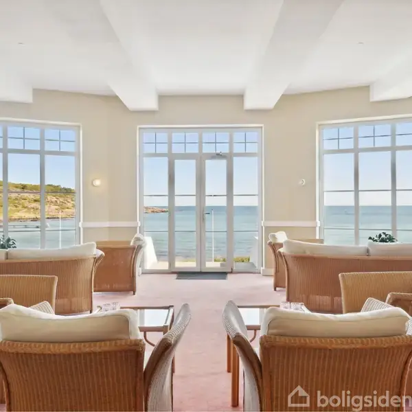 Rattanmøbler er arrangeret i en lys stue, vendt mod store vinduer med udsigt til havet og en kystlinje i det fjerne. Stemningen er rolig og stilfuld.