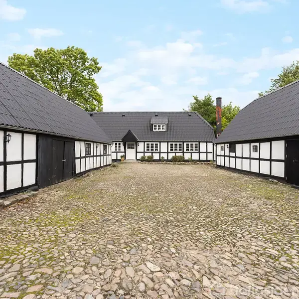 Bindingsværkshus omkranser en brostensgård. Tre bygninger danner en hesteskoformet struktur med sort og hvid facade og mørke tage. Grønt løv og blå himmel i baggrunden.