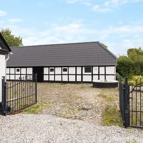 Sort-hvid bindingsværkshus står stille omgivet af grusgård og smedejernslåge. Baggrund med grønne buske og blå himmel.