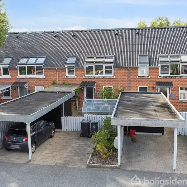 Rækkehus med røde mursten og sorte tagsten har tilknyttede carporte med parkerede biler. Omkring huset ses en betongangbro med hække og et skovområde i baggrunden.