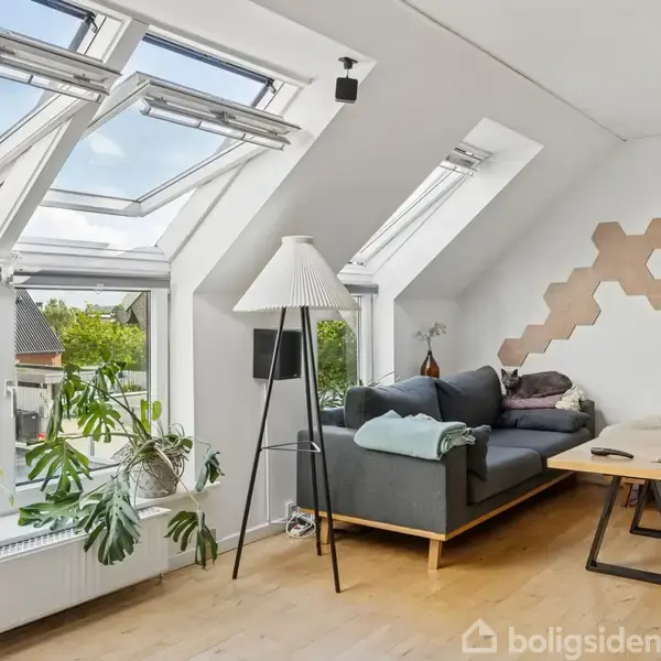 Stue med en grå sofa ved en skråvæg under loftsvinduer. Sofabord og lampe i rummet. Indendørs planter og udsigt til husenes tage og himmel udenfor. Ingen tekst.