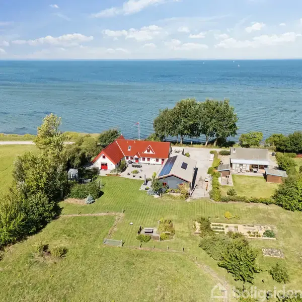 Rødt hus med en stor have, beliggende ved kysten. Havet strækker sig i baggrunden, omgivet af grønne træer og marker.