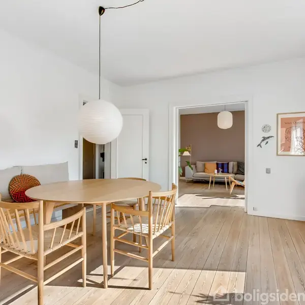 Rundt spisebord med tre stole står i en lys stue med trægulv. Et vægkunstværk ses til højre, mens en sofa er synlig i en tilstødende stue gennem åbningen.