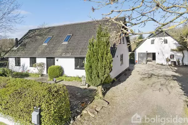 Hvidt hus med sort tag og to tagvinduer, omkranset af grønne buske og træer. Grusvej fører til en separat garagebygning i baggrunden under blå himmel.