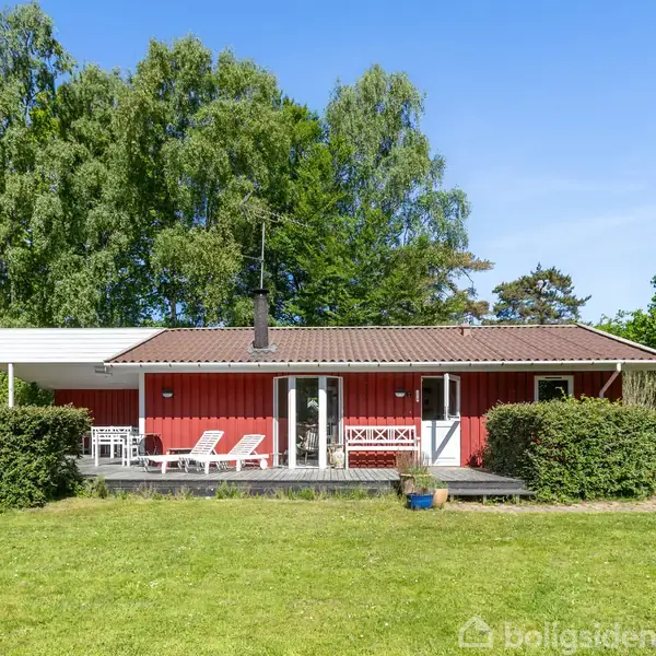 Rødt sommerhus står stille i en grøn have, omgivet af buske og store træer; to liggestole og et sæt havemøbler på terrassen under klar blå himmel.