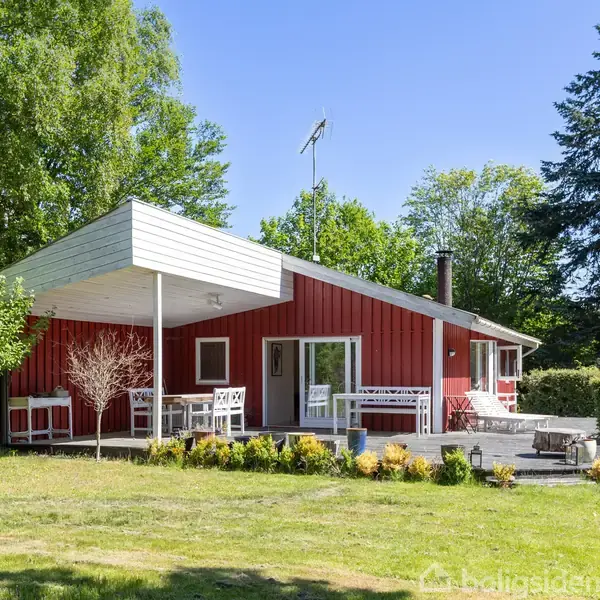 Rødt sommerhus med hvide vinduesrammer og overdækket terrasse. Havemøbler står på terrassen. Haven er grøn med høje træer og buske i baggrunden.