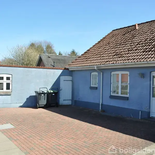 Et blåt hus med rødt tegltag står stille ved en stille vej. To affaldsbeholdere er placeret ved husmuren. Omgivet af en solrig dag og få træer.