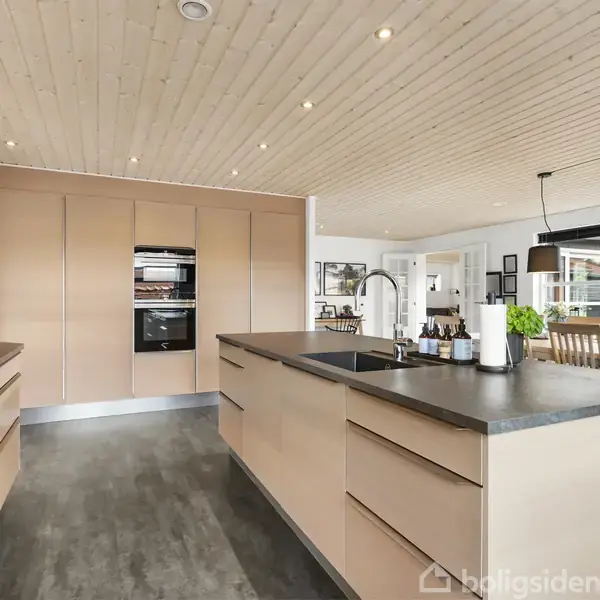 Et moderne køkken med lyse træpaneler på loftet, grå bordplader, og indbyggede skabe. En køkkenø med vask, omgivet af et spiseområde med træstole og store vinduer.