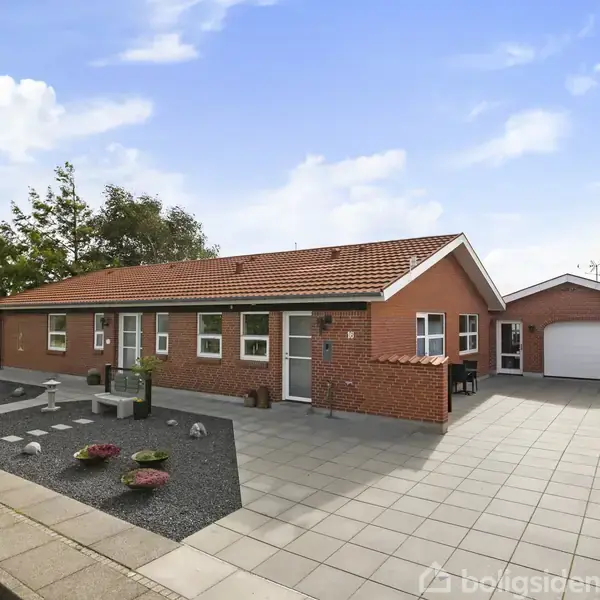 Rødt murstenshus med skråt tag står stille i et åbent, brolagt gårdmiljø. Til venstre ses grusbed med planter, mens marker og træer findes bagved.