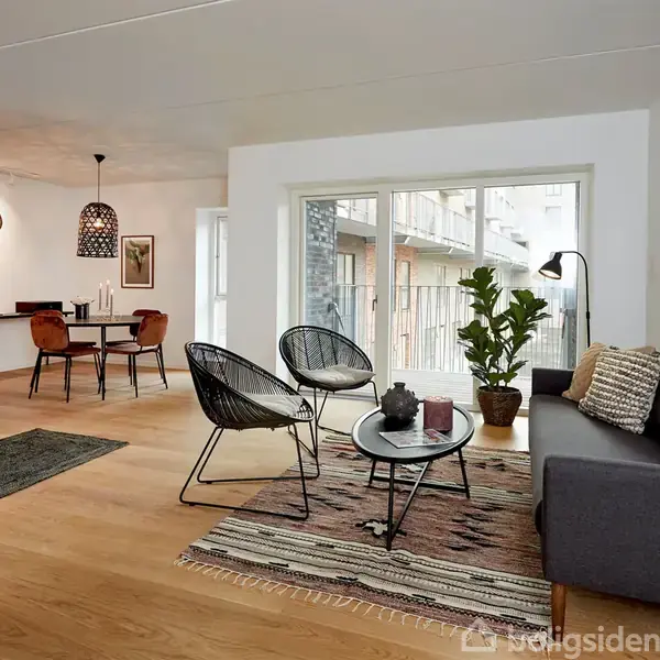 En stue med moderne indretning har en grå sofa og to sorte stole ved et lavt sofabord. Store vinduer giver udsigt til en balkon. I baggrunden ses et spisebord.