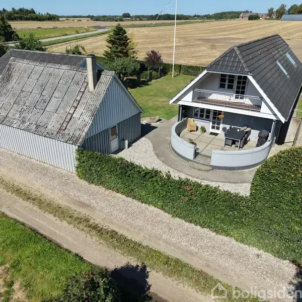 To huse står tæt ved siden af hinanden. Det ene har en terrasse med havemøbler. Omgivet af grønne hække og marker.