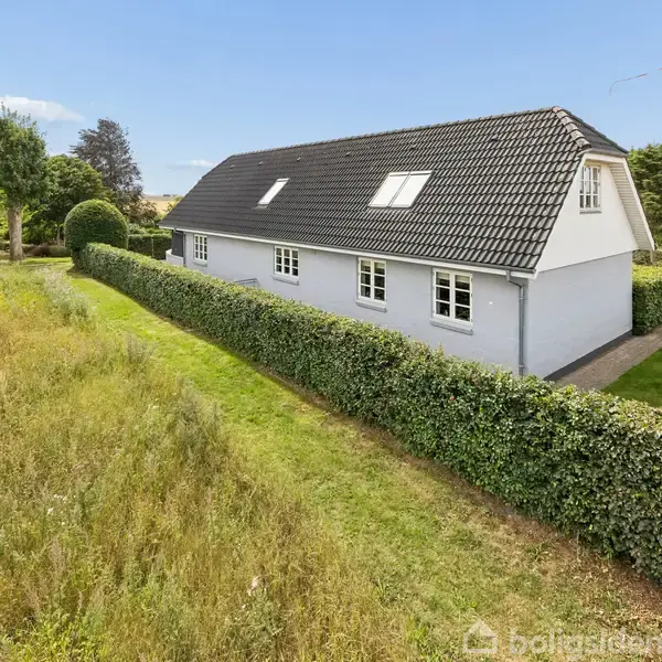 Et hvidt hus med sort tag står ved siden af en velplejet hæk. Det er omgivet af en græsplæne, og i baggrunden ses træer og en bil.