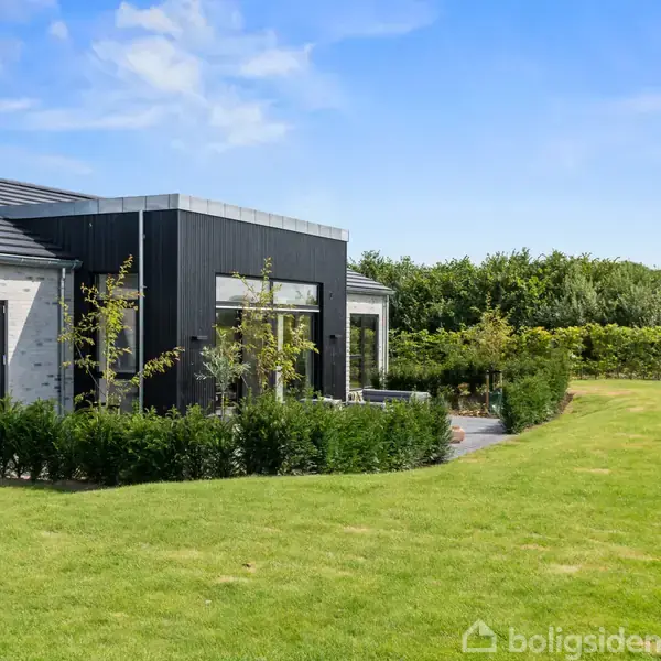 Et moderne hus med store vinduer og grå facader står i en velplejet have med grøn græsplæne og omkransende hække under en blå himmel.