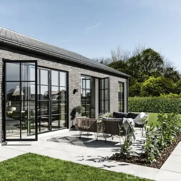 Moderne hus med store glasdøre åbner ud til en terrasse, hvor havemøbler står opstillet. Grønt græs og hække omkranser området, mens træer ses i baggrunden under en klar himmel.