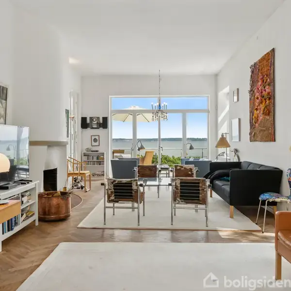 Stue med sofaer og stole omkring et bord. Store vinduer giver udsigt til havet. Bogreol, TV og skrivebord findes til venstre. Rum er lyst og moderne.