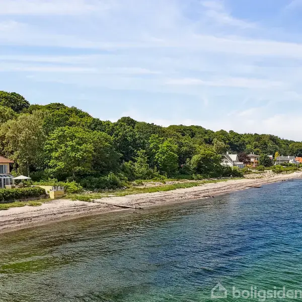 Et gult hus med rødt tag ligger ved en kyst. Det er omgivet af grønne træer. Havet strækker sig langs stranden i en fredelig landskabsindstilling.
