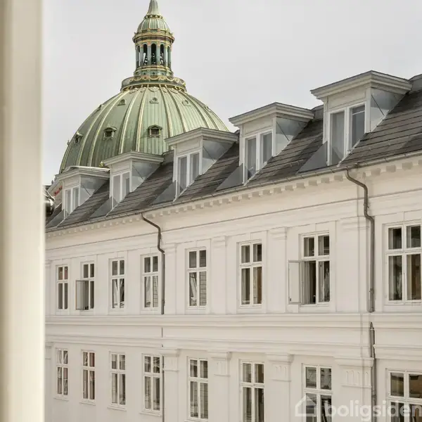 En bygning med hvide vinduer står i forgrunden, mens en stor grøn kuppel troner i baggrunden. Taget har flere små kviste med vinduer.