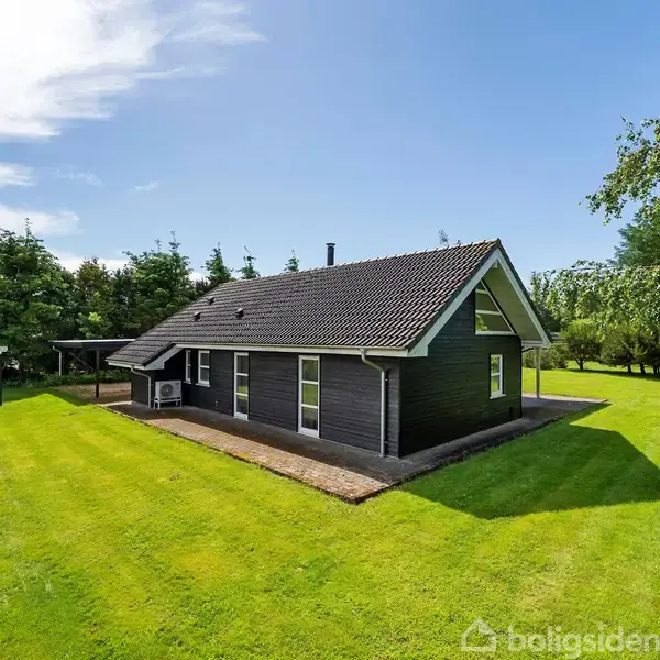Et sort træhus med skråt tag står i en solrig have med velplejede græsplæner og omkranset af træer. Terrasse med siddepladser ved siden af huset.