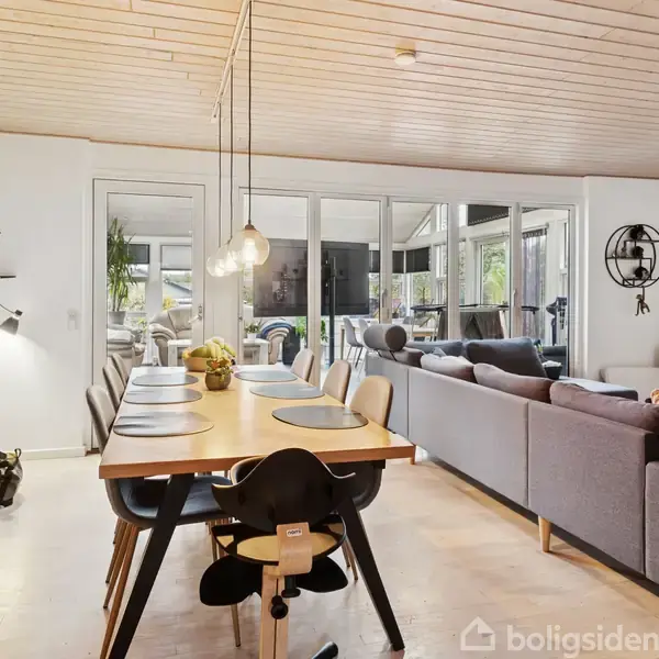 Spisebord med dækkede stole, omgivet af moderne åbent køkken-alrum med grå sofa og glasvitrineskab, i lyst rum med trægulv og -loft. Bagerst ses store vinduer og dør til terrasse.
