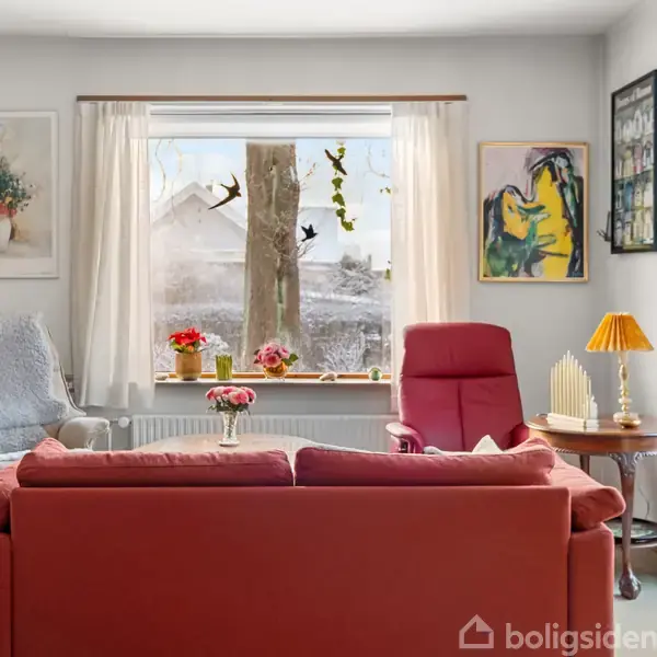 En rød sofa og to lænestole står i en lys stue med vinduesudsigt til sneklædt landskab. Væggene er dekoreret med malerier. Bordlampe med orange skærm på træbord.