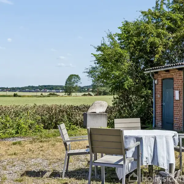 Et rundt bord med stole står placeret udendørs foran en bygning med røde mursten og mørke døre. I baggrunden ses et bredt åbent landskab under en klar blå himmel.