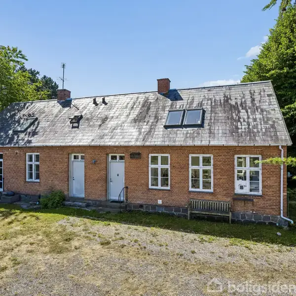Et rødt murstenshus med mørkt tag og solcelle, står i en have med træer og grusbelagt gårdsplads. Huset har flere vinduer og en hvid dør i midten.