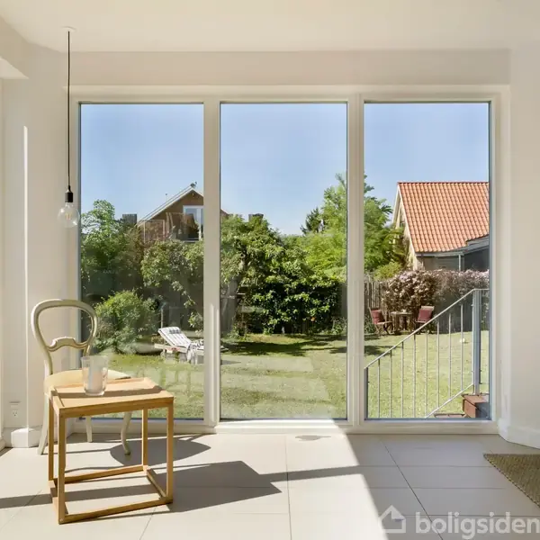 En lys stue med store vinduer, udstyret med en enkel stol og et lille bord med et glas, set mod en solrig have med grønne planter og græsplæne.
