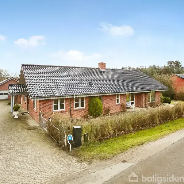 Rødt murstenshus med mørkt tag står langs vejen, flankeret af hæk og indkørsel. En bil og garage ses i baggrunden under en blå himmel.