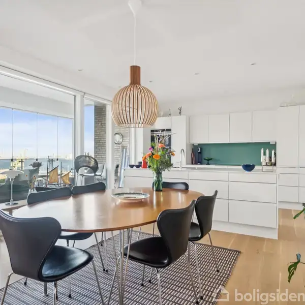 Et rundt spisebord med sorte stole står under en trælampe. I baggrunden er et moderne, lyst køkken med hvide skabe. En balkon med udsigt ses gennem store vinduer.