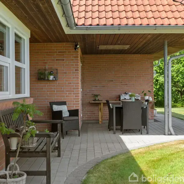 En terrasse med borde og stole ved siden af et murstenshus. Ovenover er der et tag med røde teglsten. Græsplæne og grøn have omkranser terrassen i baggrunden.