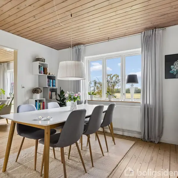 Spisebord med stole står i et lyst rum. En loftslampe hænger over bordet. Der er reoler med bøger og dekorationer. Store vinduer giver udsigt til natur.