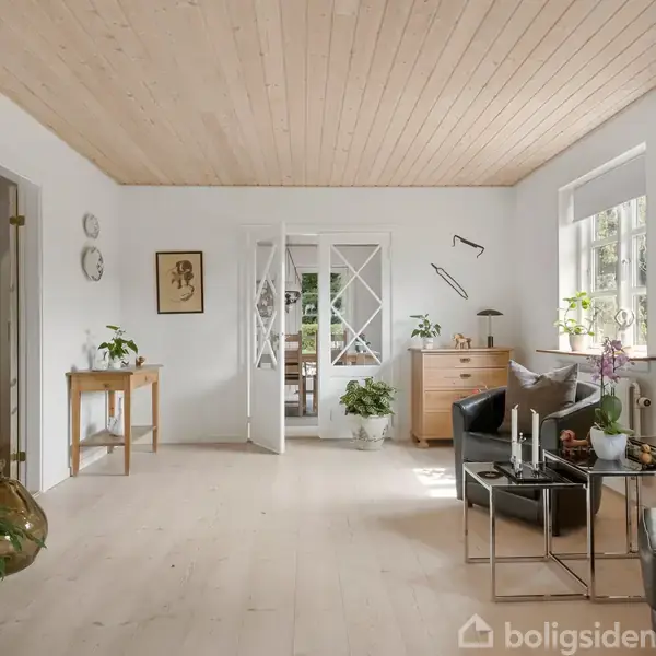 Stue indrettet med moderne møbler og planter, gennemgående lyse trælofter og -gulve. Vinduerne lader naturligt lys strømme ind. Et par sorte stole omkranser et sofabord med stearinlys.
