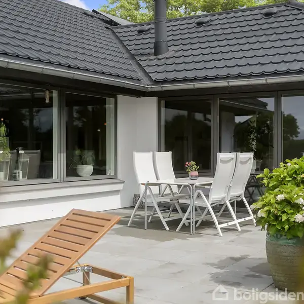 Et udendørs terrasseområde med hvide havemøbler og en solseng. Blomster i krukker står omkring bordet. Bygningen har store vinduer og et tag med sorte tegl.