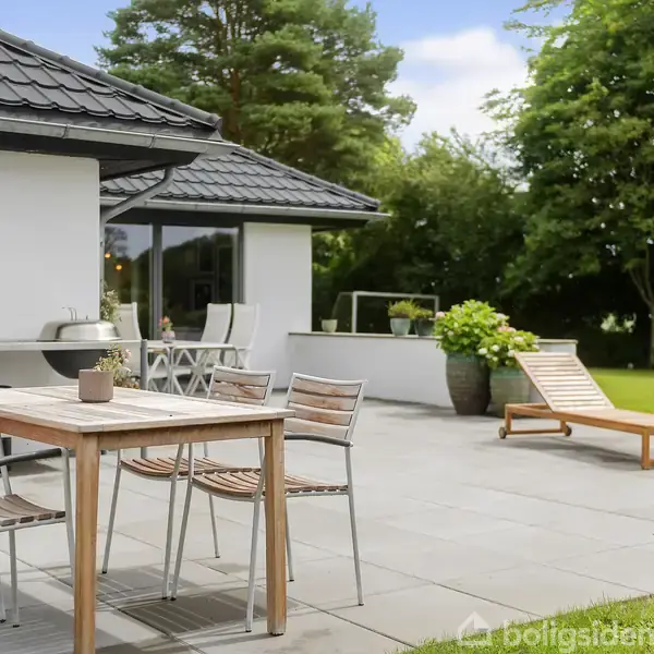 Terrassemøbler står på en flisebelagt terrasse ved et hvidt hus. Der står en grill og blomsterkrukker ved siden af med grønne træer i baggrunden.