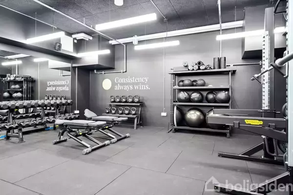 Et moderne fitnessrum med vægte og træningsbænke står klar. Sætningen "Consistency always wins." er skrevet på væggen i det lyse, velorganiserede lokale.