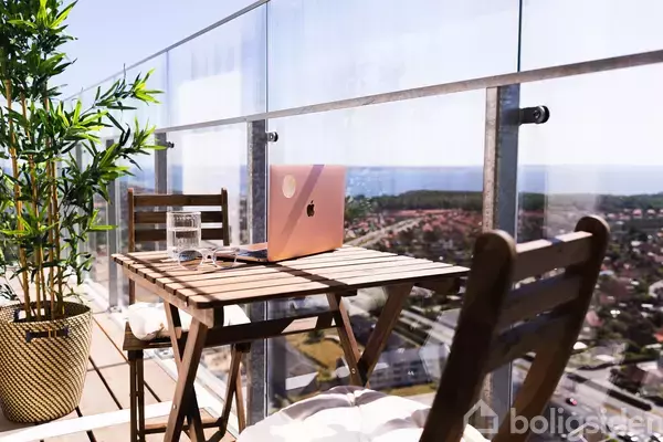 En bærbar computer står på et træbord ved en altan med transparent rækværk. Der er også et glas vand og en plante ved siden af, med udsigt over byen.
