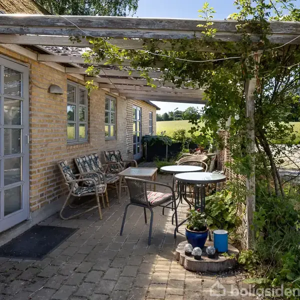 En terrasse ved et murstenshus har borde og stole under en pergola med klatrende planter. Området er omgivet af natur med udsigt til marker og åbne grønne områder.