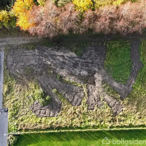 En stor skulptur af en figur ligger i græsset ved siden af en bygning. Figuren er delvist integreret i jorden omgivet af træer og grønne områder.
