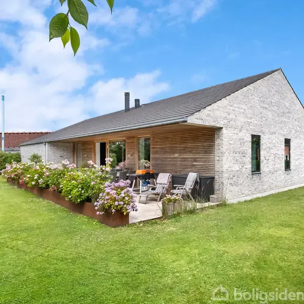 Et moderne hus i en have med blomsterbede langs terrassen. Huset har hvidt murværk, skråt tag og store vinduer. Haven er velplejet med grønne græsplæner og træer.