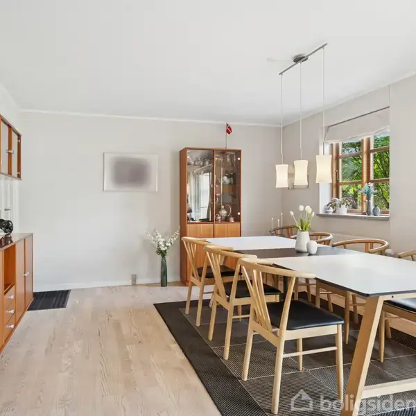 Spisebord med seks klassiske stole står på et tæppe i en lysekronebelyst spisestue. Træreoler og et vitrineskab bagved, vindue med udsigt til en grøn have.