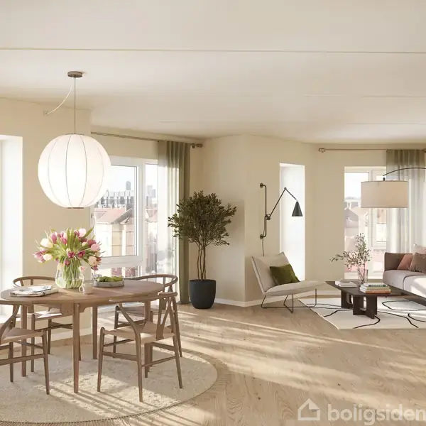 Et lyst stueområde indeholder et rundt spisebord med fire stole og en vase med blomster. En sofa med sofabord står bagved i en rummelig, elegant indrettet lejlighed med store vinduer.
