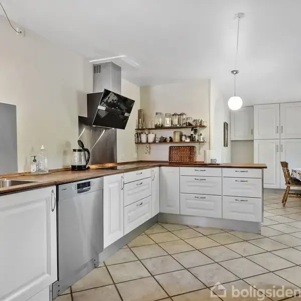 Et moderne køkken med hvide skabe og træbordplader. En indbygget ovn, opvaskemaskine og stålvask er synlige. Rummet er lyst med hvide vægge og flisegulv. Hylder med krydderier i baggrunden.