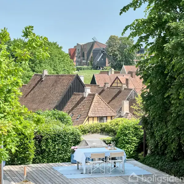 Et bord med stole står på en terrasse dækket af lys dug i en frodig have. Baggrunden viser traditionelle hustage og en moderne bygning omgivet af grønne træer.