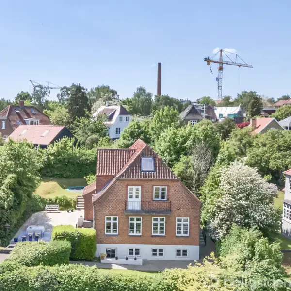 Rødt murstenshus med hvide vinduer står fredeligt blandt grønne træer i et villakvarter. Flere huse med røde tage omgiver det, og en kran ses i det fjerne under blå himmel.