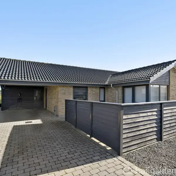 En etplans murstensvilla med sort tegltag står på en flisebelagt indkørsel. En lav, mørk træhegn adskiller forhaven fra indkørslen. Solrigt, blå himmel indikerer en klar dag.