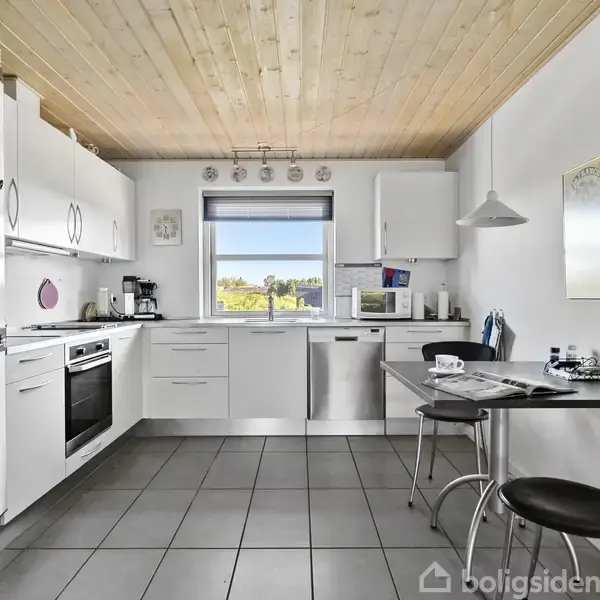 Køkken med hvide skabe og apparater, såsom opvaskemaskine og ovn. Et lille spisebord med to stole står ved siden af et vindue, der tilbyder udsigt til en grøn landskab.